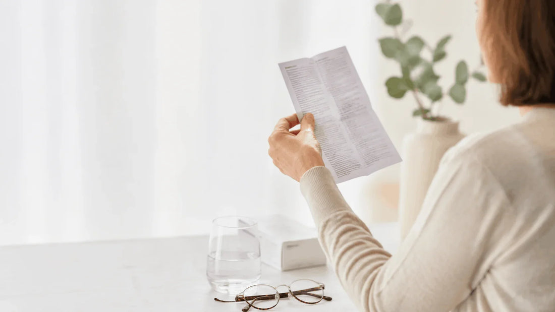 Mână ținând un prospect la distanță de citit, lângă ochelari de citit — test dioptrii acasă